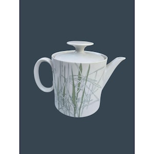 Op de afbeelding: Een witte porseleinen theepot met een cilindervormig lichaam en een bijpassend deksel met een knopgreep. De buitenkant is versierd met een patroon van dunne, overlappende groene en grijze grassprieten die rond de basis van de pot lopen.