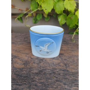 以下が含まれることがあります： 金色の縁取りのある、小さく、楕円形の青と白の磁器のカップ。カップには、青い空を背景に、白いカモメが飛んでいる様子が描かれています。