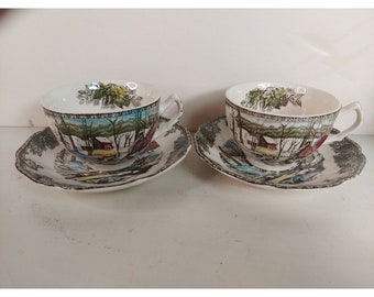Può includere: Due tazze da tè e piattini vintage con un design a tema invernale. Le tazze e i piattini sono bianchi con un motivo marrone, verde e rosso. La scena rappresenta un paesaggio innevato con un fienile rosso e alberi.