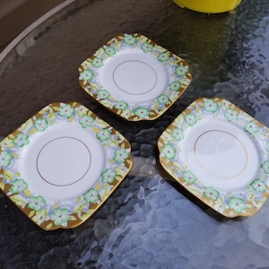 Puede incluir: Tres platos cuadrados de cerámica con un centro blanco y un diseño floral dorado y verde alrededor del borde.