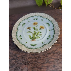 Könnte beinhalten: Eine weiße Porzellansaucers mit grünem und goldenem Rand. Die Mitte der Saucers zeigt ein florales Design mit gelben Blumen und grünen Blättern.