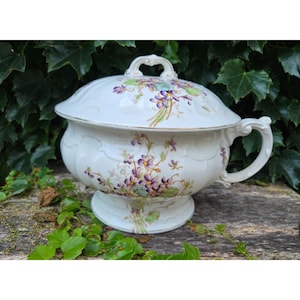 Puede incluir: Una fuente de porcelana blanca con tapa, adornada con motivos florales morados y verdes. La fuente tiene un asa lateral y un asa decorativa en la tapa. La fuente está sobre una base de pedestal.