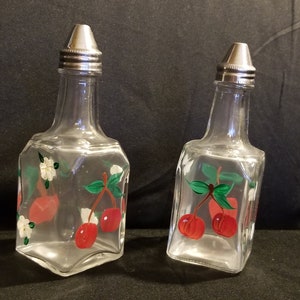 May include: Two clear glass bottles with silver tops and painted cherry designs. The bottles are on a black surface.