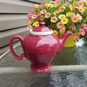 Könnte beinhalten: Eine tiefrote Teekanne mit einem weißen Deckel und einem weißen Rand am oberen Rand. Die Teekanne steht auf einer Glastischplatte.