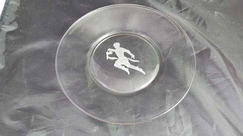 May include: A clear glass plate with an etched design of a person running. The plate is on a dark gray surface.