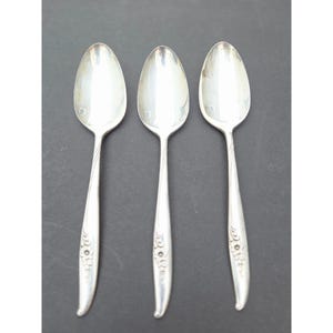 May include: Three silver spoons with oval bowls and long, slender handles. Each spoon has a small, engraved design near the base of the handle and a small square mark on the bowl. The spoons are arranged on a dark gray surface.