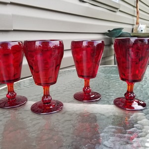 Op de afbeelding: Vier robijnrode wijnglazen met een textuurpatroon. De glazen hebben een afgeronde komvorm en een korte, amberkleurige steel. Ze zijn gerangschikt op een glazen tafel, met een neutrale achtergrond.