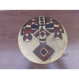 Puede incluir: Un plato redondo de cerámica beige con un diseño geométrico marrón y blanco. El diseño presenta una cara estilizada con una forma de diamante en el centro. El plato está firmado "Joyces".