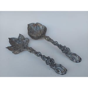May include: A set of two silver salad servers with grapevine and leaf designs. The servers are made of metal and have a vintage look.