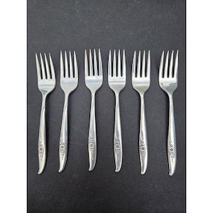 May include: Set of six silver forks arranged on a black surface. Each fork has a slender handle with a floral design near the base. The tines are evenly spaced and the forks are made of a shiny, reflective metal.