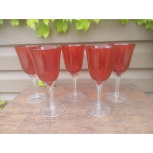 Può includere: Cinque calici da vino rossi con steli e basi trasparenti sono disposti su una superficie di legno. I calici hanno una forma a tulipano e un colore rosso vibrante, perfetti per servire il vino.