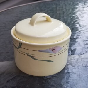 May include: A yellow ceramic sugar bowl with a lid and a floral design. The design features a stylized flower with green leaves and a gold trim.