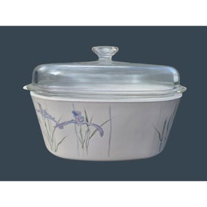 Pode incluir: Uma assadeira branca com tampa de vidro transparente. A assadeira apresenta um desenho floral de íris roxas e caules verdes. A assadeira tem aproximadamente 23 cm de largura e 33 cm de comprimento.