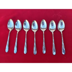 May include: Six silver teaspoons with ornate floral designs on the handles. The spoons are arranged in a row on a red surface.