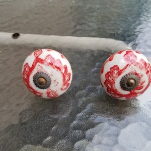 Puede incluir: Dos pomos de armario de cerámica blanca con detalles rojos y dorados. Los pomos tienen un diseño floral.