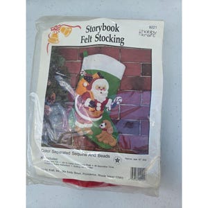 May include: A Hobby Kraft Storybook Felt Stocking craft kit in a plastic package. The packaging features a green felt stocking with a Santa Claus and puppy design, labeled 18 inches. Text includes Storybook Felt Stocking and 9221.