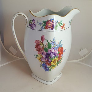 May include: A white ceramic pitcher with a gold rim and floral pattern. The pitcher has a scalloped top and a square base. The floral pattern features pink, purple, yellow, and red flowers.
