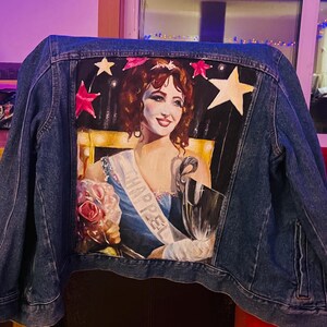 May include: A blue denim jacket with a painted image of a woman wearing a blue dress and a sash that says "CHARPEL". The woman is holding a bouquet of pink roses and a trophy. The background is black with gold stars.