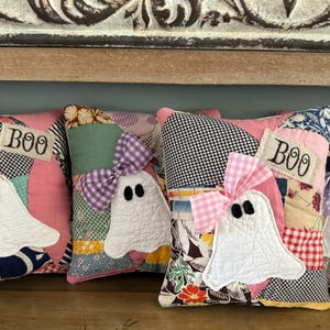 May include: Four square patchwork pillows featuring white ghost appliqués with black eyes. Each ghost wears a gingham bow in pink or purple. The pillows include fabric patches in various patterns and colors, with a rectangular tag reading BOO.