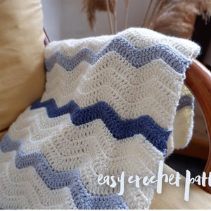 Pode incluir: Um cobertor de crochê branco e azul com um padrão chevron. O cobertor está drapeado sobre uma cadeira de madeira com uma almofada amarela. O texto "easy crochet pattern" está escrito na almofada.