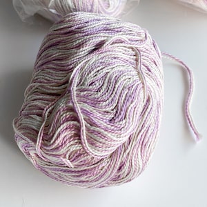 May include: A skein of white and lavender yarn with a variegated colour pattern. The yarn is in a plastic bag with text that reads "purchase sufficient yarn of same dye lot to complete garment. Hand wash separately in cold water, towel dry. Or in cold water,  scarborough, ontario, canada."