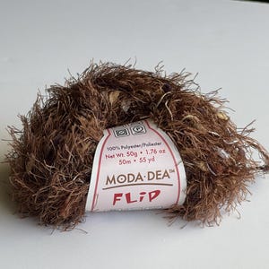 Pode incluir: Uma bola de fio marrom, bege e roxo com um rótulo branco que diz "100% Polyester/Poliester Net Wt. 50g 1.76 oz 50m 55 yd MODA DEA™ FLIP"