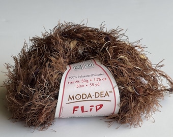以下が含まれることがあります： 茶色、ベージュ、紫色の糸の玉。白いラベルには「100% Polyester/Poliester Net Wt. 50g 1.76 oz 50m 55 yd MODA DEA™ FLIP」と書いてある