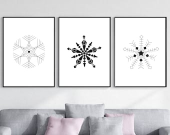 Könnte beinhalten: Drei gerahmte Kunstdrucke mit schwarz-weißen Schneeflocken-Designs. Jeder Druck zeigt ein einzigartiges Schneeflockenmuster mit geometrischen Formen und sternförmigen Zentren. Die Drucke werden über einem grauen Sofa mit rosa Kissen ausgestellt.