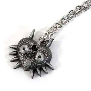 May include: A silver necklace with a pendant shaped like the Majora's Mask from the Legend of Zelda video game series.
