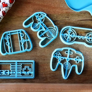 Może przedstawiać: Zestaw pięciu niebieskich plastikowych foremek do ciasteczek w kształcie różnych kontrolerów do gier wideo. Formy znajdują się na drewnianej powierzchni.