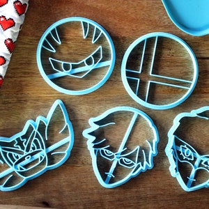 May include: Five blue plastic cookie cutters shaped like characters from a video game. The cutters are on a wooden surface.