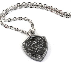 May include: A silver chain necklace with a silver pendant shaped like a shield. The shield has a black and silver design of a stylized bird with a triangle inside a circle.
