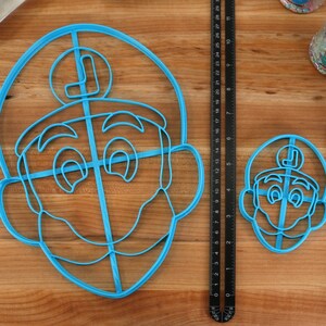 May include: Two blue plastic cookie cutters in the shape of a cartoon character's face, one large and one small. A ruler shows measurements in inches. A white oven mitt with red pixelated hearts is on the right.