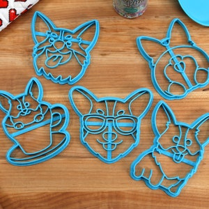 Pu&ograve; includere: Cinque tagliabiscotti in plastica blu a forma di corgi. I corgi sono in pose diverse, tra cui uno con gli occhiali da sole, uno che tiene una tazza di caff&egrave; e uno con la lingua di fuori.