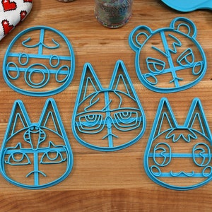 May include: Five blue plastic cookie cutters shaped like animal faces. The cutters are on a wooden surface. The animal faces include a tiger, a cat wearing glasses, a cat with a mustache, and two other cats.
