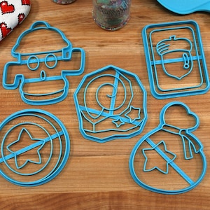 May include: Five blue plastic cookie cutters on a wooden surface. The cutters are shaped like a fire hydrant, a bag of coins, a star, an acorn, and a swirl with stars.