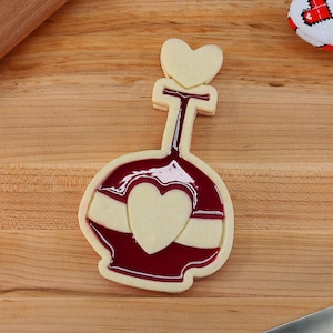 May include: A white sugar cookie shaped like a bottle with a red heart design. The cookie is decorated with red icing and a white heart.