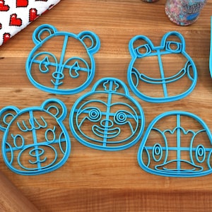 May include: Five blue plastic cookie cutters shaped like animal faces. The cutters are on a wooden surface. The animals are a panda, a frog, a sloth, a bear, and a penguin.