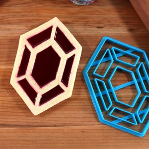 Op de afbeelding: Een blauwe plastic koekjesvormer in de vorm van een geometrische zeshoek met een kleinere zeshoek erin. Ernaast ligt een koekje met een vergelijkbare vorm gevuld met rode gelei.