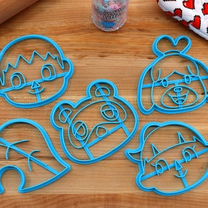 May include: Five blue plastic cookie cutters shaped like characters from the video game Animal Crossing: New Horizons. The characters are Tom Nook, Isabelle, K.K. Slider, Marshal, and a leaf.
