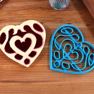 May include: A blue heart-shaped cookie cutter with a detailed design and a heart-shaped cookie with a red filling.