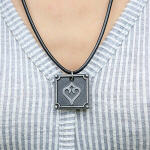 May include: A silver pendant necklace with a square pendant featuring a stylized heart design. The pendant is on a black cord.