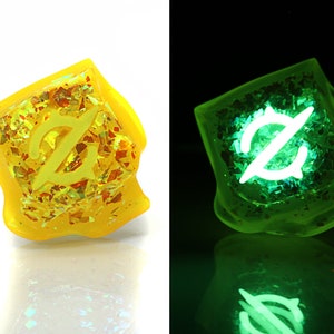 May include: A yellow dice with a white symbol on it. The dice is made of resin and has a glittery finish. The dice glows green in the dark.