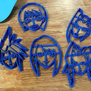 May include: Five blue plastic cookie cutters shaped like cartoon characters with different hairstyles and facial expressions.