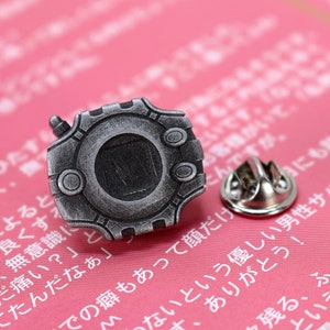 May include: A silver-colored metal pin shaped like a video game device with a black screen and a small antenna. The pin has a safety clasp on the back.