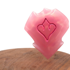 May include: A pink, shield-shaped decorative item with a heart-shaped design carved into its surface. The item is displayed on a wooden surface against a white background. The shield has a glossy finish.