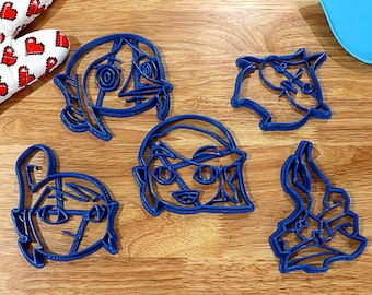Puede incluir: Cinco cortadores de galletas de plástico azul con forma de caras de dibujos animados. Los cortadores están sobre una superficie de madera.