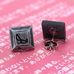 May include: A pair of silver earrings with a square design. The earrings feature a black design on the front. The earrings are displayed on a pink surface.