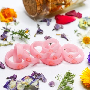 May include: A set of six pink silicone rings with a heart design. The rings are stacked on top of each other and are surrounded by dried flowers.