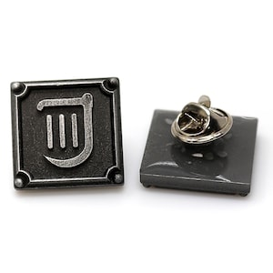 Op de afbeelding: Een zilveren metalen pin met een zwarte achtergrond en een zilveren harp symbool. De pin heeft een metalen sluiting aan de achterkant.
